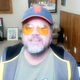 Kyanka sits in front of a computer, wearing yellow sunglasses and a baseball cap. Next to him is a keyboard.